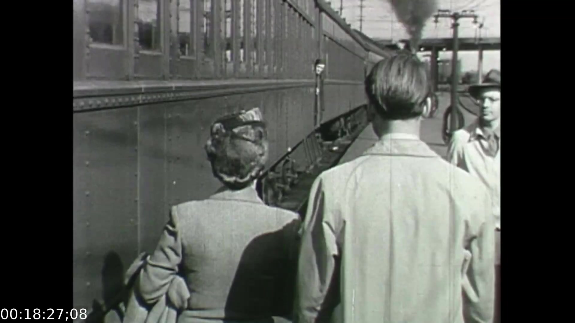 The still from 'Feelings of Depression' (1950) captures a moment at a train station, showcasing two figures—a man and a woman—standing side by side, gazing at a train in the background. The woman, dressed in a vintage outfit with a decorative hat, appears to embody a mid-20th-century aesthetic, while the man is seen from behind, likely in a lighter coat. The scene reflects a time of travel and transition, paralleling the film's exploration of emotional struggles, particularly in the context of the protagonist John Murray's experiences with depression. The historical setting, characterized by the steam train, emphasizes the era's social dynamics and the significance of mental health awareness during the period.