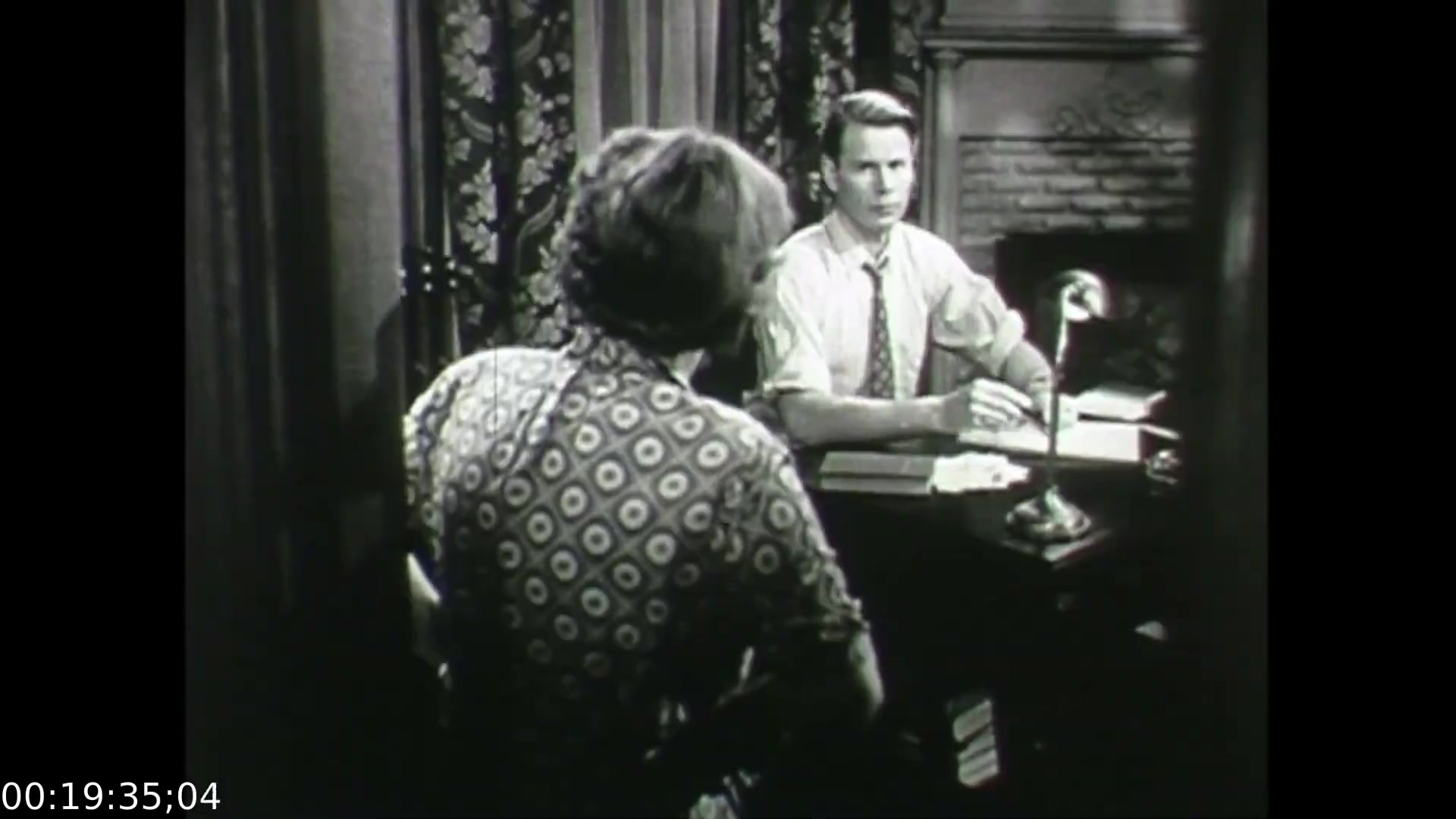 The still from 'Feelings of Depression' (1950) depicts a pivotal moment in the film, featuring a tense conversation between two characters. One is seated at a desk, appearing engaged and serious, while the other, partially obscured from view, seems introspective. The setting suggests a formal environment, likely a consultation scene, characterized by period-appropriate decor and attire. This frame encapsulates the film's focus on mental health, illustrating the struggles of John Murray and the interpersonal dynamics of discussing depression.