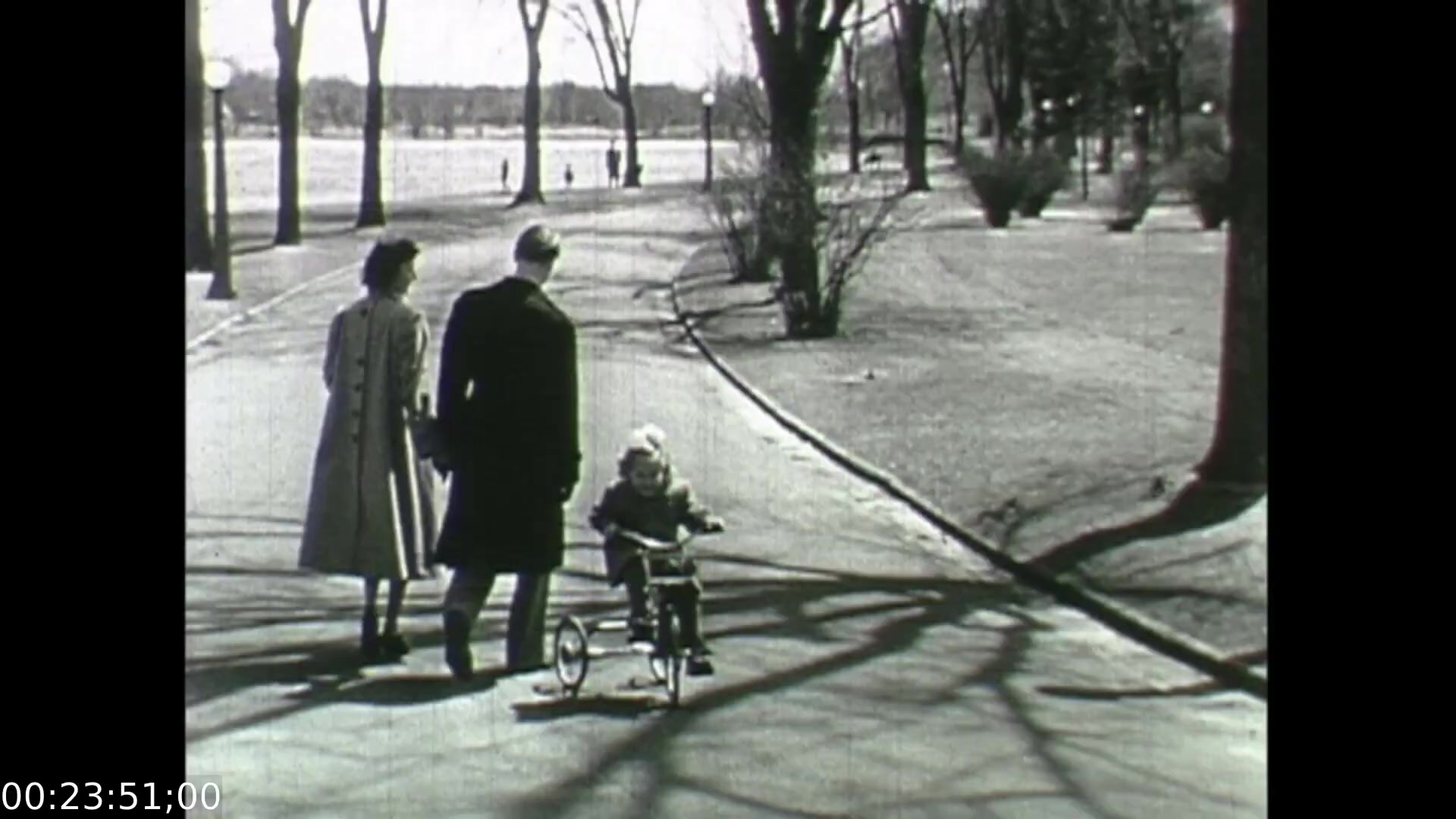 The still from 'Feelings of Depression' (1950) depicts a serene park scene with three figures: a man and woman walking hand-in-hand while watching a child on a tricycle. Shadows from the trees stretch across the pathway, enhancing the tranquil yet somber atmosphere. This moment emphasizes familial connection and innocence, contrasting with the film's exploration of mental health struggles faced by the central character, John Murray. The stark black-and-white imagery reflects the film's post-war context and highlights societal attitudes towards depression during that era.