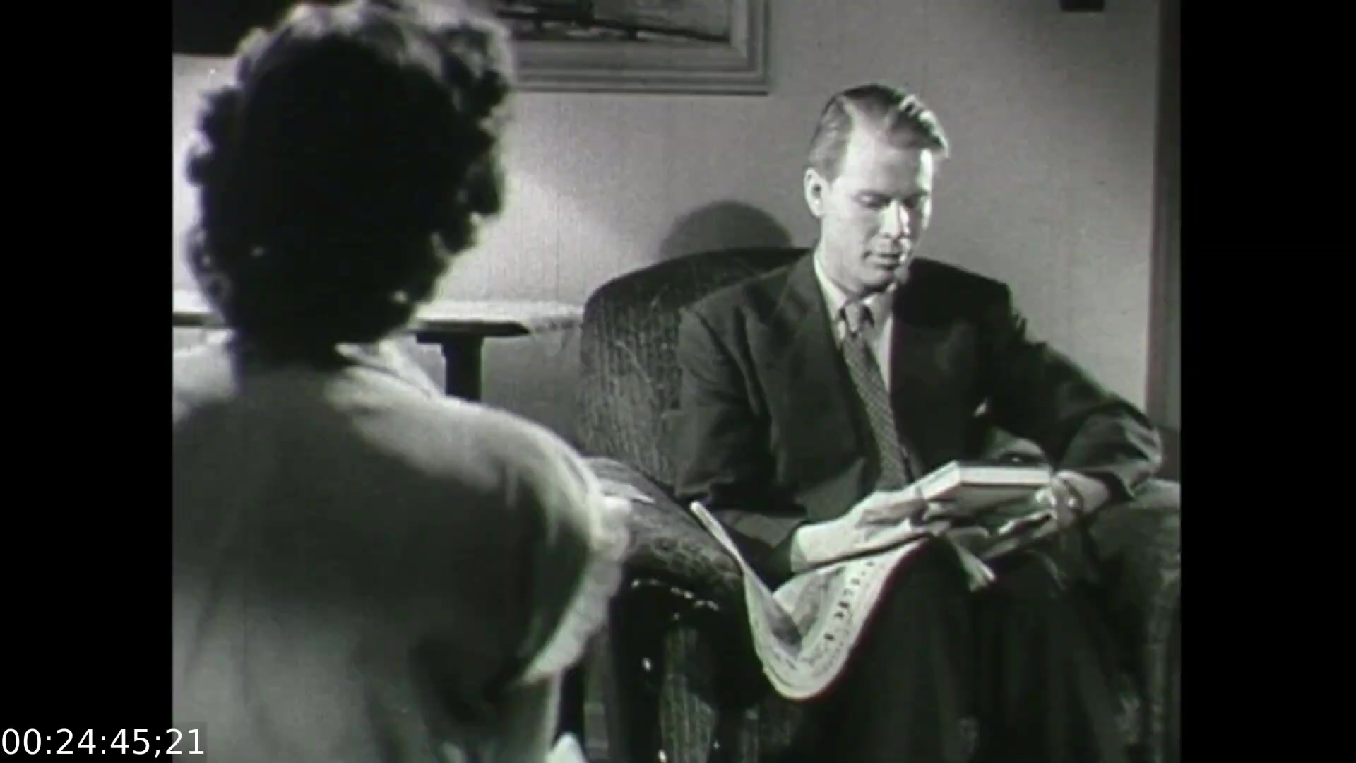 The still from 'Feelings of Depression' (1950) depicts a tense moment in a counseling session. A businessman, John Murray, sits in a chair, visibly absorbed and possibly troubled as he looks at a book in his hands. His serious demeanor suggests he is grappling with emotional issues. In front of him, a woman, likely a therapist or confidante, is turned slightly away, her back facing the camera. This setup emphasizes the intimate yet difficult nature of their conversation, highlighting themes of despondency and the search for understanding in the postwar context of mental health.