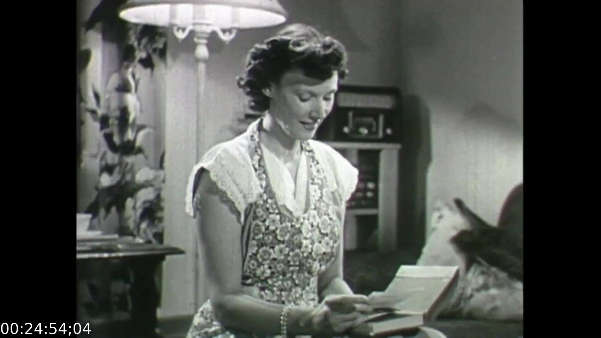 The still from 'Feelings of Depression' depicts a young woman seated in a cozy, domestic setting, engaged in reading a letter. She wears a floral apron, suggesting a homemaker role, and her expression hints at a moment of contemplation or emotional significance in her reading. The background features a lamp and a radio, contributing to a warm, intimate atmosphere. This scene likely underscores the film's exploration of personal feelings and relationships in the context of depression, reflecting social dynamics of the era.
