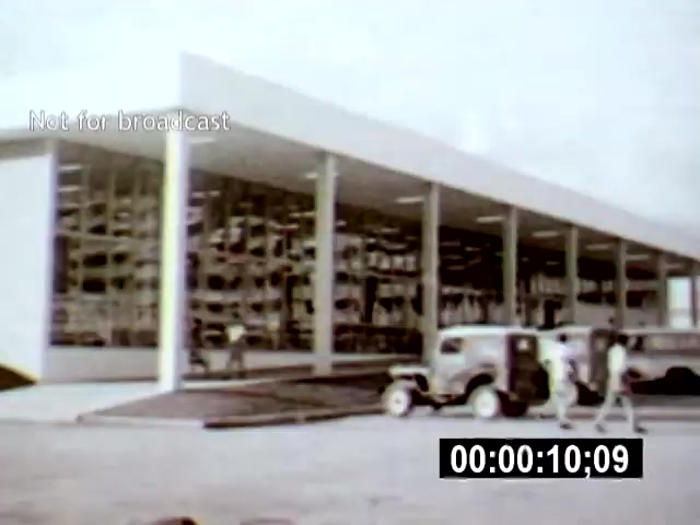 The film still depicts a modernist building in Brasília, characterized by its clean lines and expansive glass facade. The camera angle shows the structure from a slight distance, capturing both the building and individuals walking nearby, which emphasizes the scale of the architecture in relation to human activity.