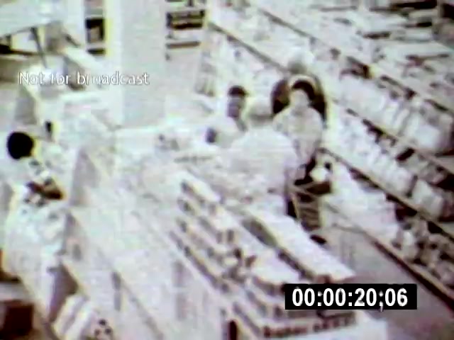 The still from 'Brazilia' captures a busy supermarket scene, focusing on multiple individuals engaging in conversation among rows of products. The overhead camera angle emphasizes the spaciousness of the setting, highlighting the organized chaos of consumerism.