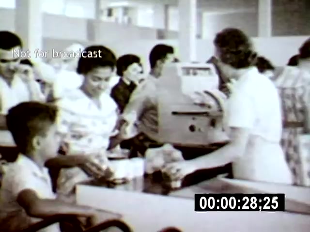 The film still depicts a busy grocery store scene in Brasília, featuring a diverse group of people interacting at the checkout counters. Shot from a slightly elevated angle, the focus is on a woman assisting customers, with a young boy and others visible, capturing the bustling atmosphere of daily life in the city.