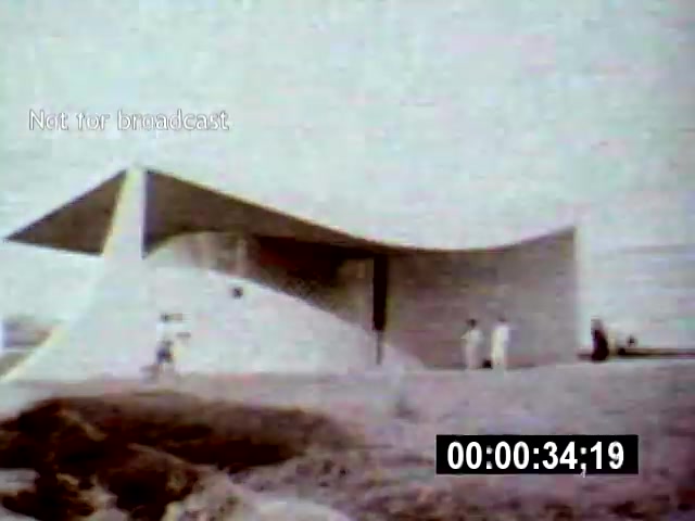 The still captures a striking modernist architectural structure in Brasília, characterized by its smooth, curvilinear forms. Captured from a low angle, a few figures are seen interacting in the foreground, emphasizing the scale and design of the building against the stark landscape.