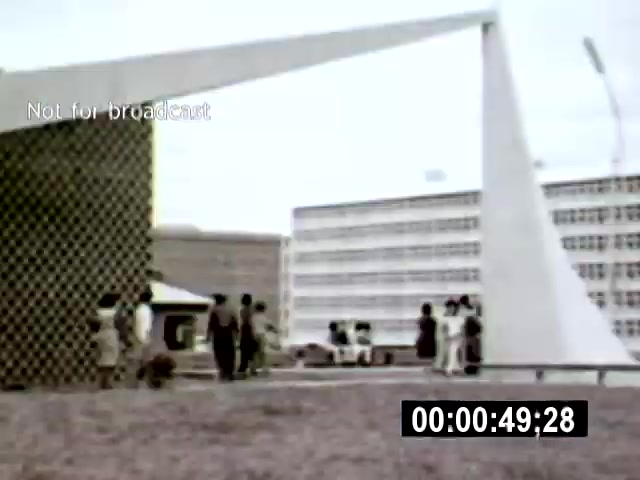 The film still from 'Brasilia' features a modernist architectural setting with a striking geometric structure in the foreground, surrounded by a group of people. The camera angle captures the scene from a low vantage point, emphasizing the dynamic lines of the architecture against a backdrop of stark buildings and a cloudy sky.