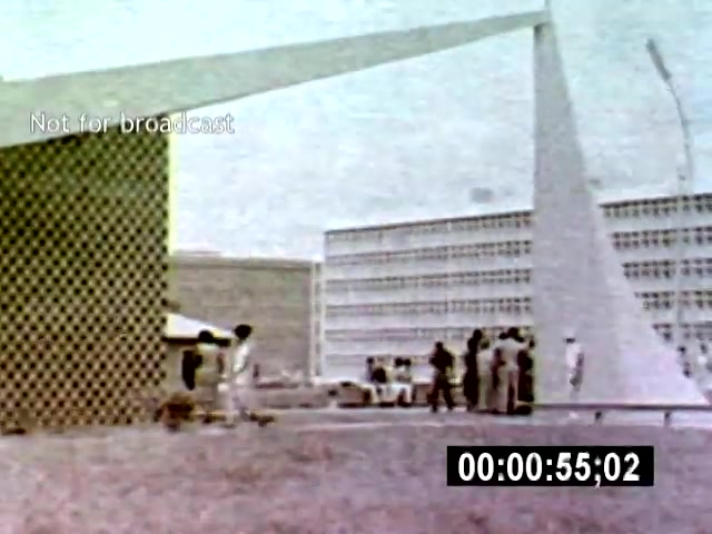 The film still captures a group of people gathering near a modern, geometric structure in Brasília, with distinctive elongated architectural features in the backdrop. The camera angle is slightly elevated, emphasizing the dynamic interaction between the subjects and the sleek lines of the buildings surrounding them.