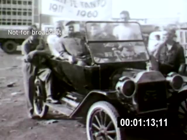 The still features a group of men gathered around a vintage car in a dusty setting, likely in Brasília. The camera angle captures the subjects from a side perspective, emphasizing their interactions with the vehicle against the backdrop of signage that hints at historical context.