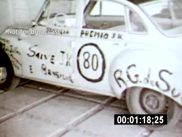The film still features a vintage car adorned with graffiti-like messages, prominently displaying '80' and other phrases celebrating 'JK' (Juscelino Kubitschek). The camera angle captures the vehicle from the side, set against a backdrop of a sparse, industrial environment, which may suggest themes of celebration amidst an evolving urban landscape in Brasília.