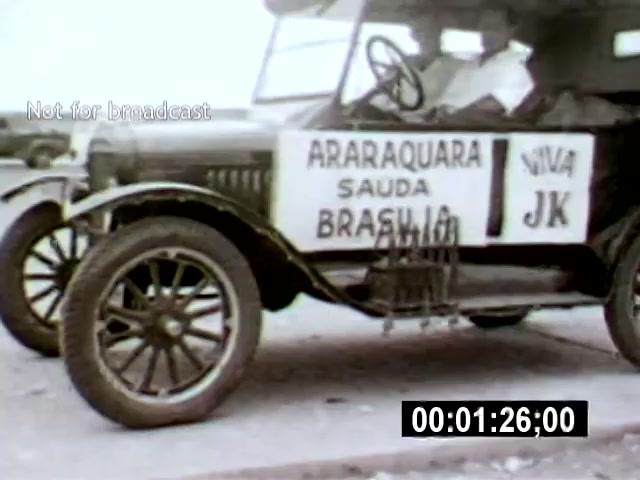 The still captures an early automobile adorned with signs saying 'ARARAQUARA SAUDA BRASÍLIA' and 'VIVA JK,' suggesting a celebratory or welcoming context related to Brasília. The camera angle is positioned to emphasize the front and side of the car, highlighting its vintage design against a blurred background.