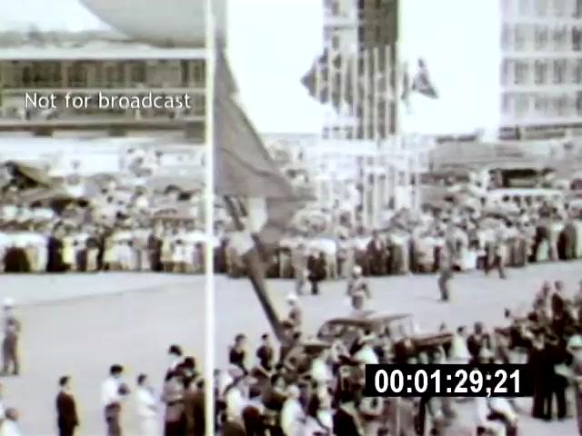The 16mm film still from 'Brasilia' captures a large gathering of people in a bustling urban setting, likely during a significant event. The camera angle offers a wide view of the crowd, focusing on the flag and the surrounding architecture, conveying a sense of civic pride and communal spirit.