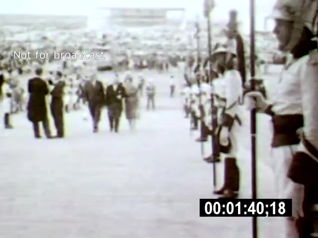 The still shows a ceremonial scene in Brasília, featuring soldiers in uniform standing in formation along a pathway, while a group of well-dressed individuals walks toward the camera in the background. The shot is taken from a low angle, emphasizing the soldiers' presence and the grandeur of the event, with a large crowd visible in the blurred background.