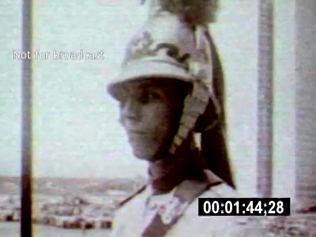 The still features a soldier in a traditional military uniform and helmet, standing at attention, likely overlooking a cityscape that suggests the modern architecture of Brasília. The camera angle captures him in close-up, emphasizing the soldier's expression and the historical context of the setting.