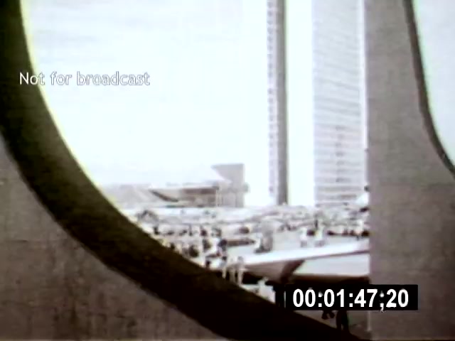 The still features an architectural view of Brasília, framed by a circular opening, showcasing the city's modernist structures and a bustling scene below. The camera angle emphasizes the contrast between the bold geometric forms and the activity of the people, highlighting the urban landscape's dynamic nature.