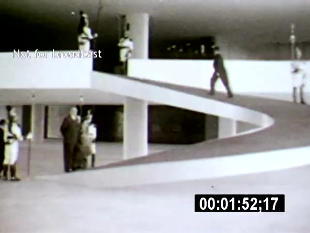 The film still from 'Brasilia' showcases a sleek, modern architectural interior with minimalist design elements, featuring individuals in formal attire interacting beneath a sweeping, curved walkway. The camera angle captures both the upward and downward perspectives, emphasizing the expansive, open space characteristic of the city’s design.
