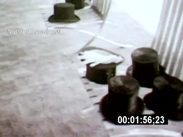 The film still from 'Brazilia' features a series of black top hats arranged on the ground, hinting at a surreal or whimsical atmosphere. The low camera angle emphasizes the hats' presence against a minimalist, light background, contributing to an intriguing sense of depth and perspective.