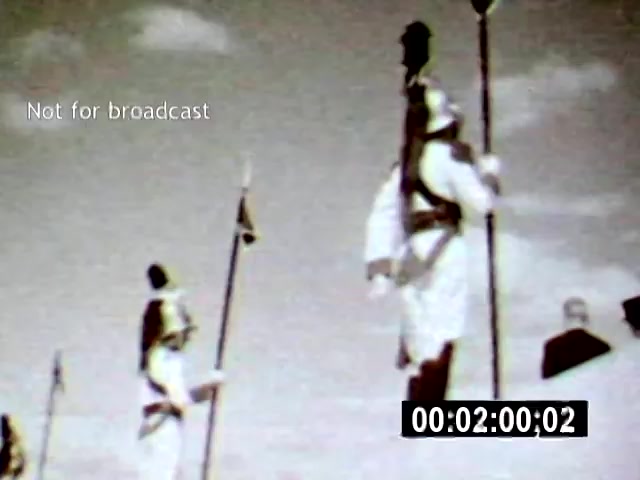 The still from 'Brazilia' depicts figures in traditional attire, likely representing cultural or historical aspects of Brazil. The camera angle suggests a slightly elevated view, capturing the subjects as they stand with poles, set against a cloudy sky, enhancing a sense of solemnity or tradition.