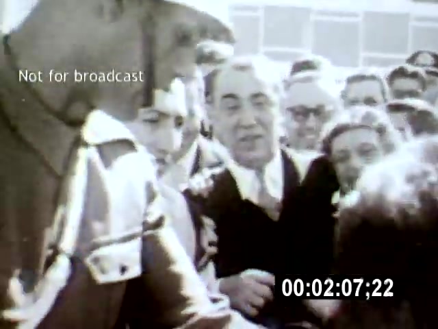 The film still from 'Brasilia' captures a crowded scene where a uniformed soldier interacts with a group of onlookers, including a prominent man in a suit. The camera angle is close, focusing on the subjects' expressions and the intensity of the moment amidst the modern architectural backdrop of Brasília.