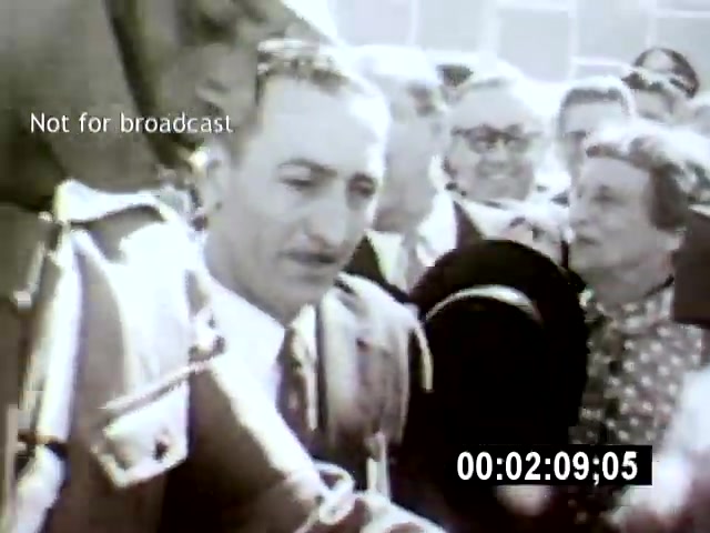 The film still from 'Brazilia' captures a moment of a crowd interacting with a man in a suit, likely an important figure, amidst a backdrop of onlookers in contemporary attire. The camera angle is close-up, emphasizing the subject's expressions and the engagement of the surrounding crowd.