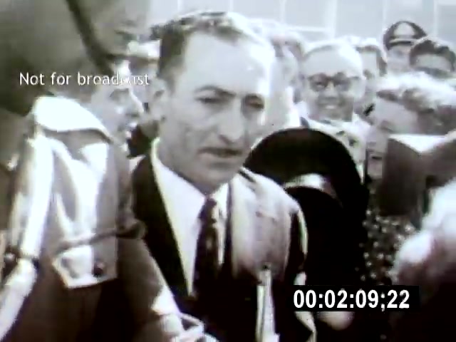 The 16mm film still from 'Brasilia' captures a group of people engaged in a moment of interaction, focusing on a well-dressed man, likely an important figure, surrounded by onlookers. The camera angle suggests a close-up perspective, emphasizing the crowd's excitement or curiosity in a bustling setting.