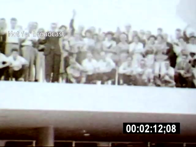 The film still displays a large group of people gathered on a balcony, looking towards the camera, with one individual prominently raising their hand. The setting appears to be a public event in Brasília, captured from a slightly lower camera angle to emphasize the crowd's engagement and the architectural backdrop.