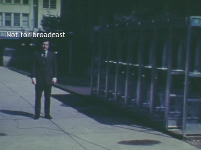 The film still depicts a man in a suit standing in an urban setting, possibly near a bus station or public transit area, with a row of telephone booths visible in the background. The camera angle is slightly low, emphasizing the subject against the backdrop of a bustling city environment.