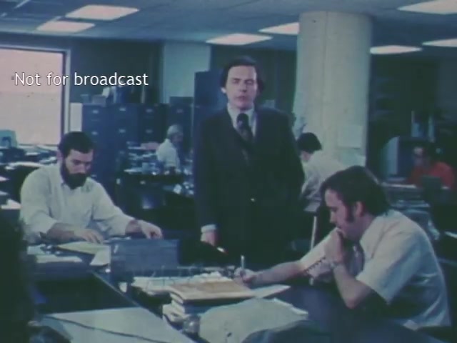 The still depicts an office setting with several men engaged in various tasks, highlighting a central figure in a suit who stands and addresses the camera. The environment is bustling yet focused, indicative of a 1970s workplace atmosphere.
