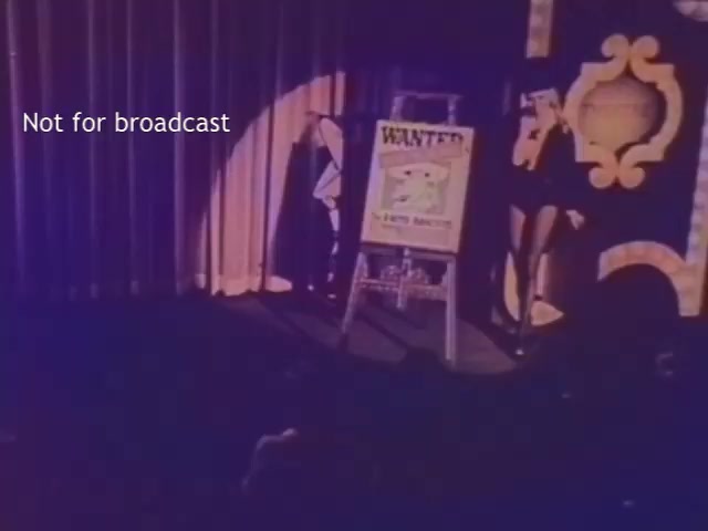 The still features a stage scene with dramatic lighting and curtains. A 'WANTED' poster is prominently displayed on an easel, featuring cartoon imagery of a character. Two figures, partially obscured, stand to the sides of the poster. The visual quality reflects the colorization process by Magix Vegas 19, indicating an older film format. A watermark notes that the image is not for broadcast.